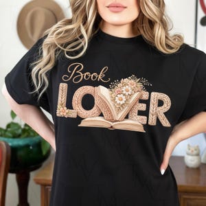 Peut inclure: T-shirt noir avec l'inscription "Book Lover" dans une police décorative. Le mot "LOVER" est conçu pour ressembler à du fil tricoté, avec un arrangement floral au centre. Un livre ouvert avec des fleurs et des feuilles est également présent.