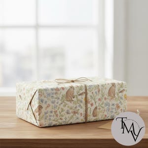 Peut inclure: Un coffret cadeau rectangulaire enveloppé dans du papier à motifs floraux avec des lapins, des oiseaux et des fleurs aux couleurs pastel. La boîte est attachée avec de la ficelle et repose sur une surface en bois.