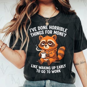 Peut inclure: T-shirt noir avec le texte "I'VE DONE HORRIBLE THINGS FOR MONEY LIKE WAKING UP EARLY TO GO TO WORK." Le motif présente un raton laveur de dessin animé en sweat à capuche orange, tenant une tasse de café.