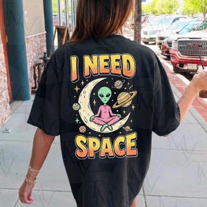 Peut inclure: T-shirt noir avec l'inscription "I NEED SPACE" en orange et jaune. Le motif représente un extraterrestre vert méditant sur un croissant de lune, entouré de planètes et d'étoiles. L'extraterrestre porte une tenue rose.