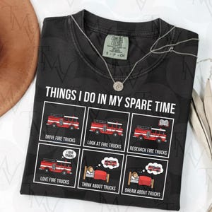 Peut inclure: T-shirt noir avec le texte "THINGS I DO IN MY SPARE TIME" et des illustrations de camions de pompiers. Le t-shirt comprend des phrases comme "DRIVE FIRE TRUCKS" et "DREAM ABOUT TRUCKS". Un collier argenté est drapé sur le t-shirt.