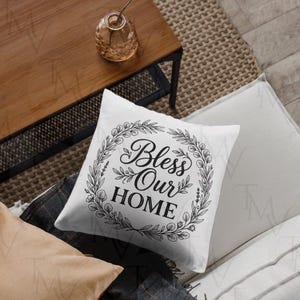Peut inclure: Un coussin carré blanc avec un motif imprimé noir représentant une couronne circulaire faite de feuilles et de tiges. À l'intérieur de la couronne, le texte indique Bless Our HOME dans une combinaison de typographie cursive et serif.