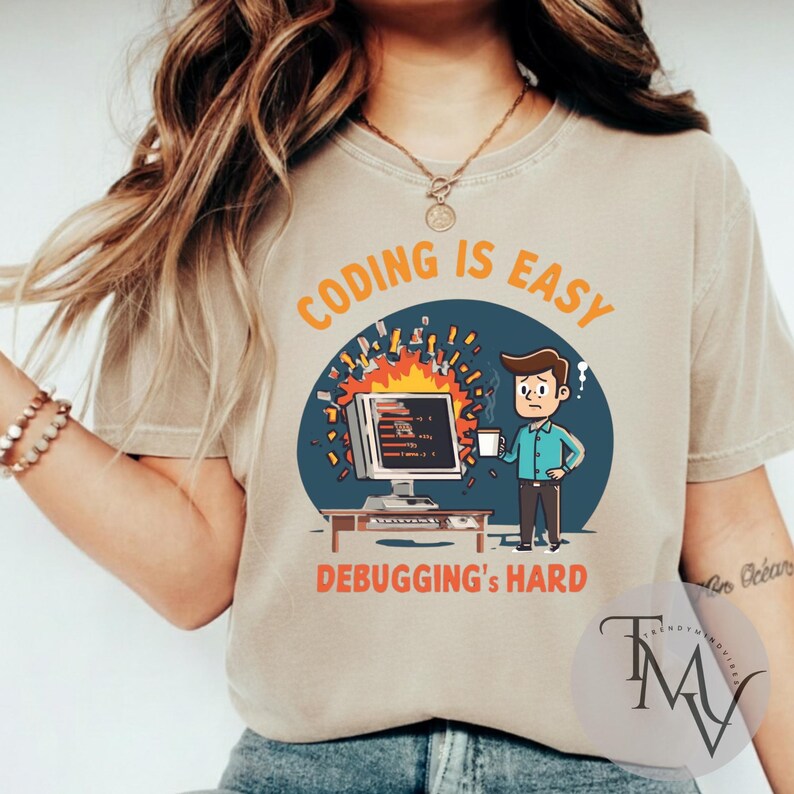 Op de afbeelding: Beige t-shirt met de tekst "CODING IS EASY" in oranje en "DEBUGGING'S HARD" in rood. De afbeelding toont een computer in brand, een persoon met een mok en een bureau. Een casual kledingstuk.