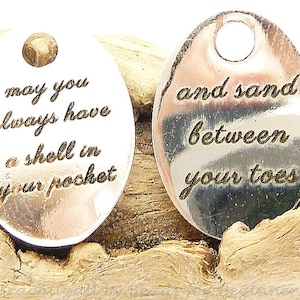 May include: Two silver oval-shaped charms with engraved text. The first charm reads "may you always have a shell in your pocket". The second charm reads "and sand between your toes".