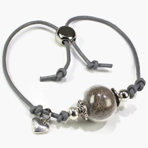 Può includere: Un braccialetto in pelle grigia con un ciondolo a forma di cuore argentato e una perla di vetro trasparente riempita di pelliccia marrone. Il braccialetto ha una chiusura a nodo scorrevole.