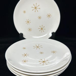 May include: A set of white ceramic plates with a gold snowflake design. The plates are round and have a simple, elegant aesthetic. The image shows a stack of four plates and one plate on a black stand.