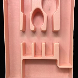 May include: A rectangular, light peach-colored plastic cutlery tray featuring molded compartments for organizing silverware. The tray includes a long vertical slot, a rounded spoon-shaped section, and several smaller rectangular dividers.