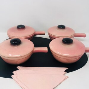 May include: Four pink ceramic ramekins with black handles and lids. The ramekins are on a black surface with a pink napkin.