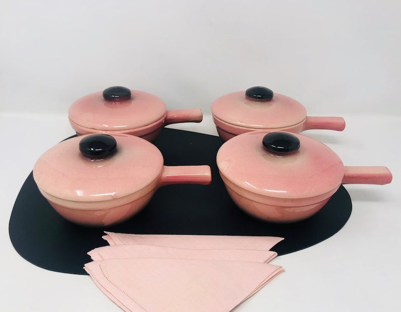 May include: Four pink ceramic ramekins with black handles and lids. The ramekins are on a black surface with a pink napkin.