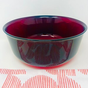 May include: A deep red glass bowl with a dark rim. The bowl has a smooth, glossy finish and a rounded shape. The bowl is sitting on a white surface with a red and white patterned cloth.