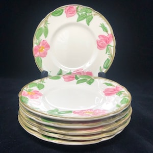 May include: A stack of six white ceramic plates with a floral design featuring pink roses and green leaves. The plates have a scalloped edge and a gold trim.