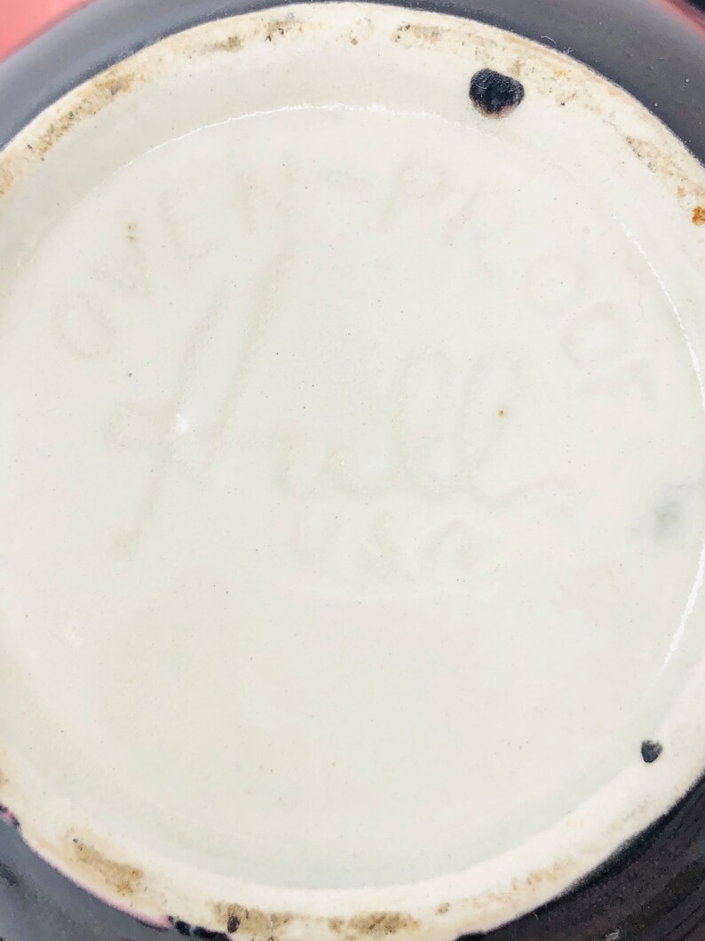 May include: Close-up of the base of a ceramic vase with a black glaze. The base is marked with the text "S.F. Hale" in black.