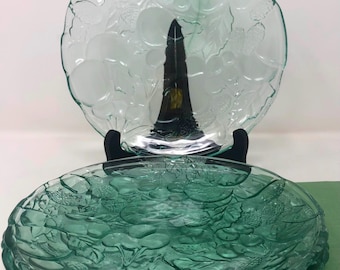 May include: Two clear glass plates with embossed fruit designs. The plates are a light green color and have a scalloped edge. One plate is on a black stand. The plates are likely for serving or display.