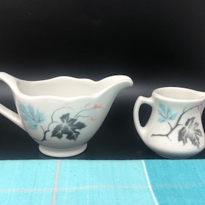 May include: Two white ceramic creamer pitchers with a vine and leaf design in shades of blue, gray, and pink. The larger pitcher has a wide, rounded base and a spout. The smaller pitcher has a more traditional shape.