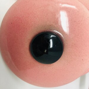 May include: A pink ceramic lid with a black circular knob in the centre. The lid has a glossy finish and a speckled texture.