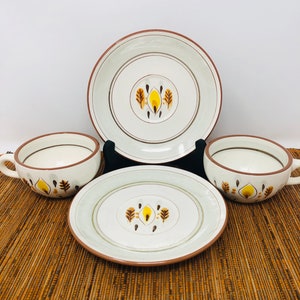 May include: A set of four ceramic dishes: two plates and two cups. The plates and cups are white with a brown rim and a central design of yellow and brown leaf-like shapes. The set is arranged on a brown woven mat.