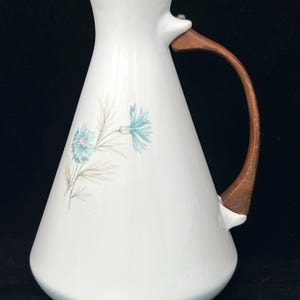 May include: A white ceramic pitcher with a flared base and a curved wooden handle. The body of the pitcher features a delicate botanical illustration of two light blue flowers with thin green stems and leaves on the front surface.