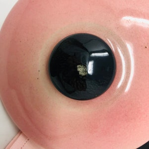 May include: A pink ceramic lid with a black knob in the centre. The lid has a slight sheen and a smooth surface.