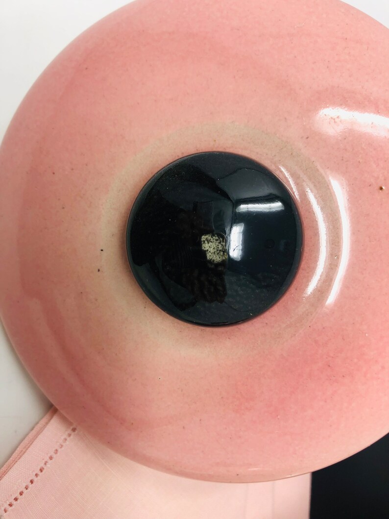May include: A pink ceramic lid with a black knob in the centre. The lid has a slight sheen and a smooth surface.