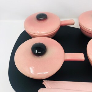 May include: Set of four pink ceramic ramekins with black lids and handles. The ramekins are on a black tray with a pink napkin.