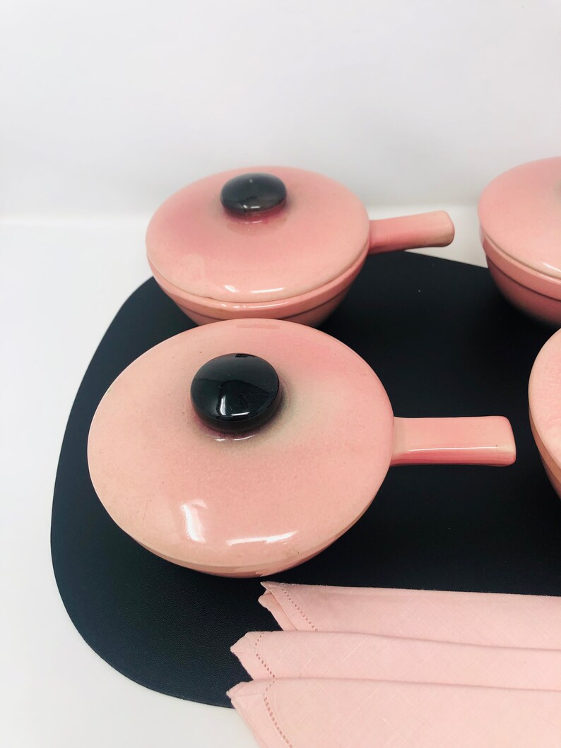May include: Set of four pink ceramic ramekins with black lids and handles. The ramekins are on a black tray with a pink napkin.