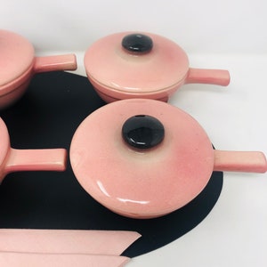 May include: Set of four pink ceramic individual serving dishes with lids and black handles. Each dish has a black knob on the lid.