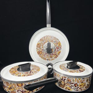 May include: A set of three white cookware pieces featuring a detailed brown, yellow, and orange floral paisley pattern. The set includes two pots with black handles and lids, and one large skillet with a long black handle and a matching lid.