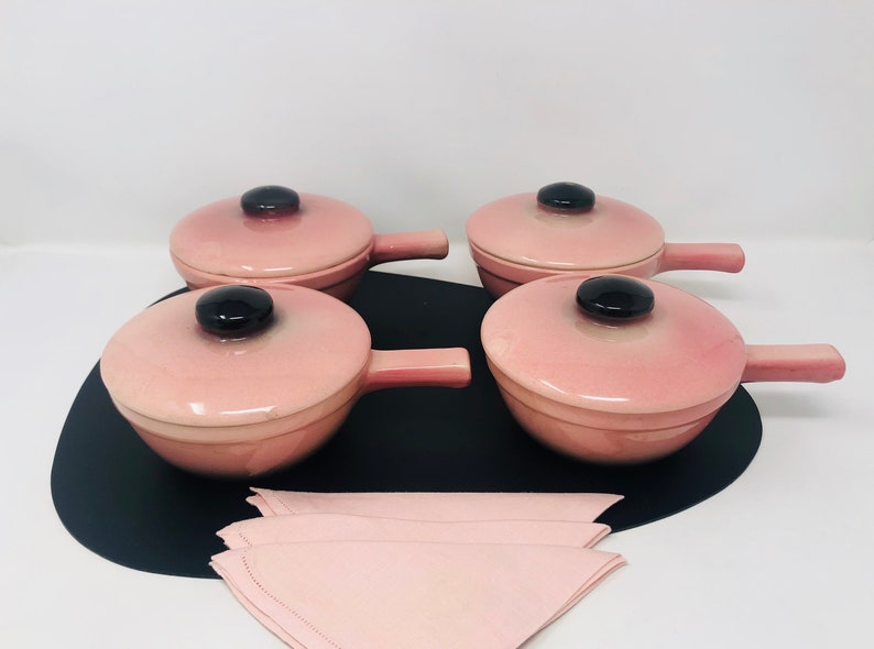 May include: Set of four pink ceramic ramekins with black handles and lids. The ramekins are on a black tray with a pink napkin.