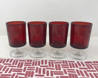 May include: Four clear glass stemmed wine glasses with a deep red hue. The glasses are arranged in a row on a white and red patterned surface.