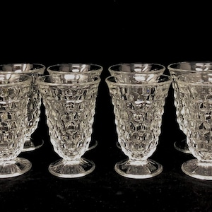 May include: A set of eight clear glass tumblers featuring a textured, geometric hobnail pattern across the exterior. Each tumbler has a flared rim, a tapered body, and a circular pedestal base, providing a consistent design across all pieces.