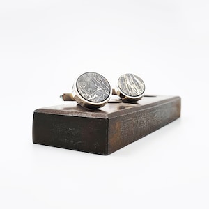 May include: A pair of silver cufflinks with a dark grey, marbled design, displayed on a wooden stand.