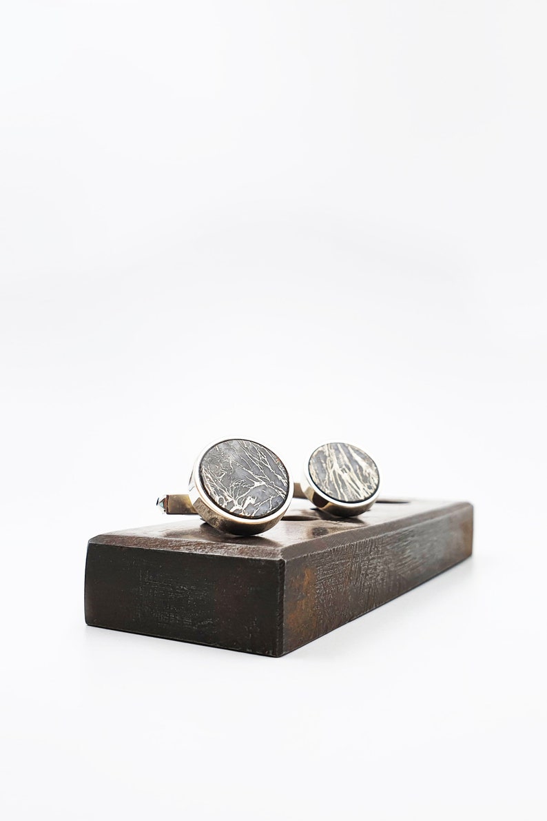 Puede incluir: Un par de gemelos de plata con un dise&ntilde;o de m&aacute;rmol gris oscuro, exhibidos en un soporte de madera.