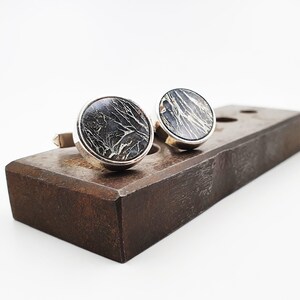 Puede incluir: Un par de gemelos de plata con inserciones redondas de m&aacute;rmol negro y dorado. Los gemelos se muestran sobre una superficie de madera.