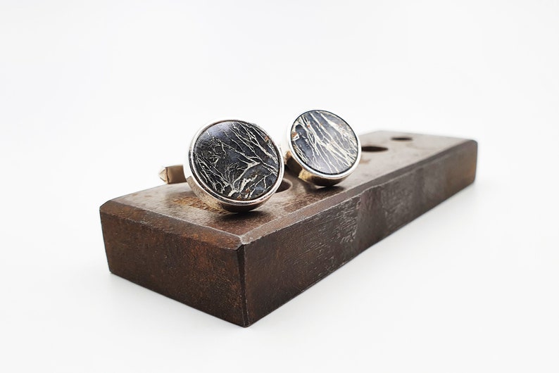Puede incluir: Un par de gemelos de plata con inserciones redondas de m&aacute;rmol negro y dorado. Los gemelos se muestran sobre una superficie de madera.