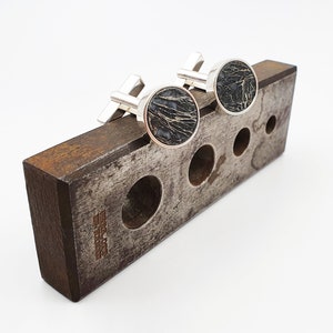 Puede incluir: Un par de gemelos de plata con incrustaciones circulares negras en un expositor de madera.