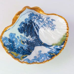 May include: A decorative seashell dish with a gold-painted rim. The interior features a blue and white wave design, reminiscent of Japanese art. The dish is ideal for holding small items.
