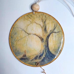 May include: A circular hanging ornament featuring a printed illustration of a fox sitting beneath a large, sprawling tree. The artwork uses soft yellow, green, and brown tones. The disc has a gold-painted edge, a wooden bead at the top, and a small metal charm.