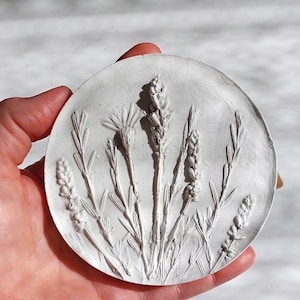 May include: A circular white clay plaque featuring a raised botanical relief of various wildflowers and lavender sprigs. The textured surface displays intricate details of stems, leaves, and flower buds in a monochromatic white finish.