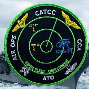 May include: A black and green circular patch with white text that reads "AIR OPS CATCC CCA FIFTH FLEET DEPLOYMENT ATO" and a radar screen with a green line pointing to the right. The patch also features a yellow airplane wing with a white star on each side.