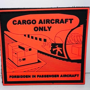 May include: A square patch with a black border and background. The patch features an orange design with the words "CARGO AIRCRAFT ONLY" above an illustration of a cargo plane and boxes. The text "FORBIDDEN IN PASSENGER AIRCRAFT" is below the illustration.