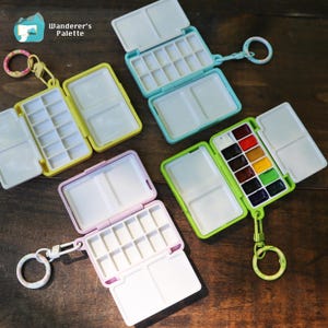 May include: Four portable watercolor palettes with keychains, featuring yellow, light blue, lavender, and lime green plastic cases. Each palette includes multiple white mixing wells and paint compartments, with one set containing twelve small paint pans.