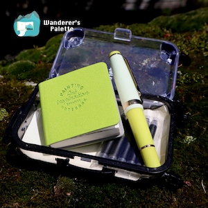 May include: A lime green notebook with the words "Painting Art Inspiration Notebook" is open inside a clear plastic case. A yellow and white pen and ink cartridges are also in the case. The case is on a bed of green moss.