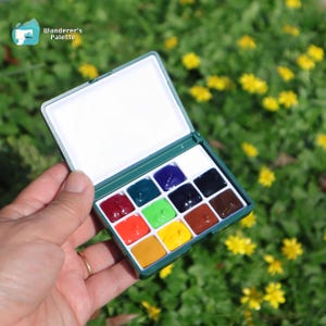 May include: A green watercolor paint set with 12 vibrant colors, held open against a backdrop of green foliage and yellow flowers. The set includes a white mixing area and is labeled "Wanderer's Palette".