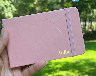 May include: A dusty rose-colored notebook with an elastic band closure. The notebook has a geometric design on the cover and the name "Julia" in gold lettering. The upper right corner has the text "Wanderer's Palette".
