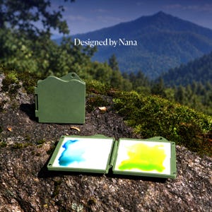 May include: A portable olive green watercolor palette case with a hinged design. One side is closed, while the other is open, revealing two white mixing wells containing blue and yellow paint. The text "Designed by Nana" is printed above the palette.