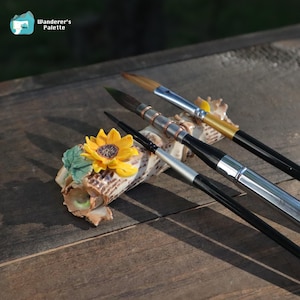 May include: Four paint brushes with black handles and silver or gold ferrules are arranged on a decorative holder with a sunflower and leaves. The holder is made of a light brown material. The logo "Wanderer's Palette" is in the upper left corner.