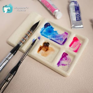May include: A ceramic watercolor palette with blue, purple, pink, and brown paint. Two paintbrushes and two tubes of watercolor paint are also visible. The palette has the text "Wanderer's Palette" and a logo in the upper left corner.