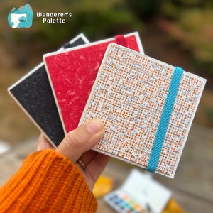 May include: A hand holds three small square notebooks with textured covers in black, red, and patterned white. The white notebook has a blue elastic band. The notebooks are from Wanderer's Palette.