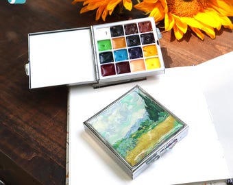 May include: A metal watercolor paint set featuring a square case with a landscape painting on the lid and an open interior with sixteen individual paint pans in various colors. The set is displayed alongside a closed case and a blank sketchbook.