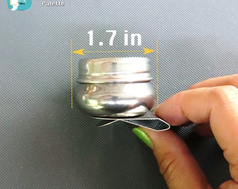 May include: A silver metal container with a lid, measuring 1.7 inches in height. The container is being held by a hand.
